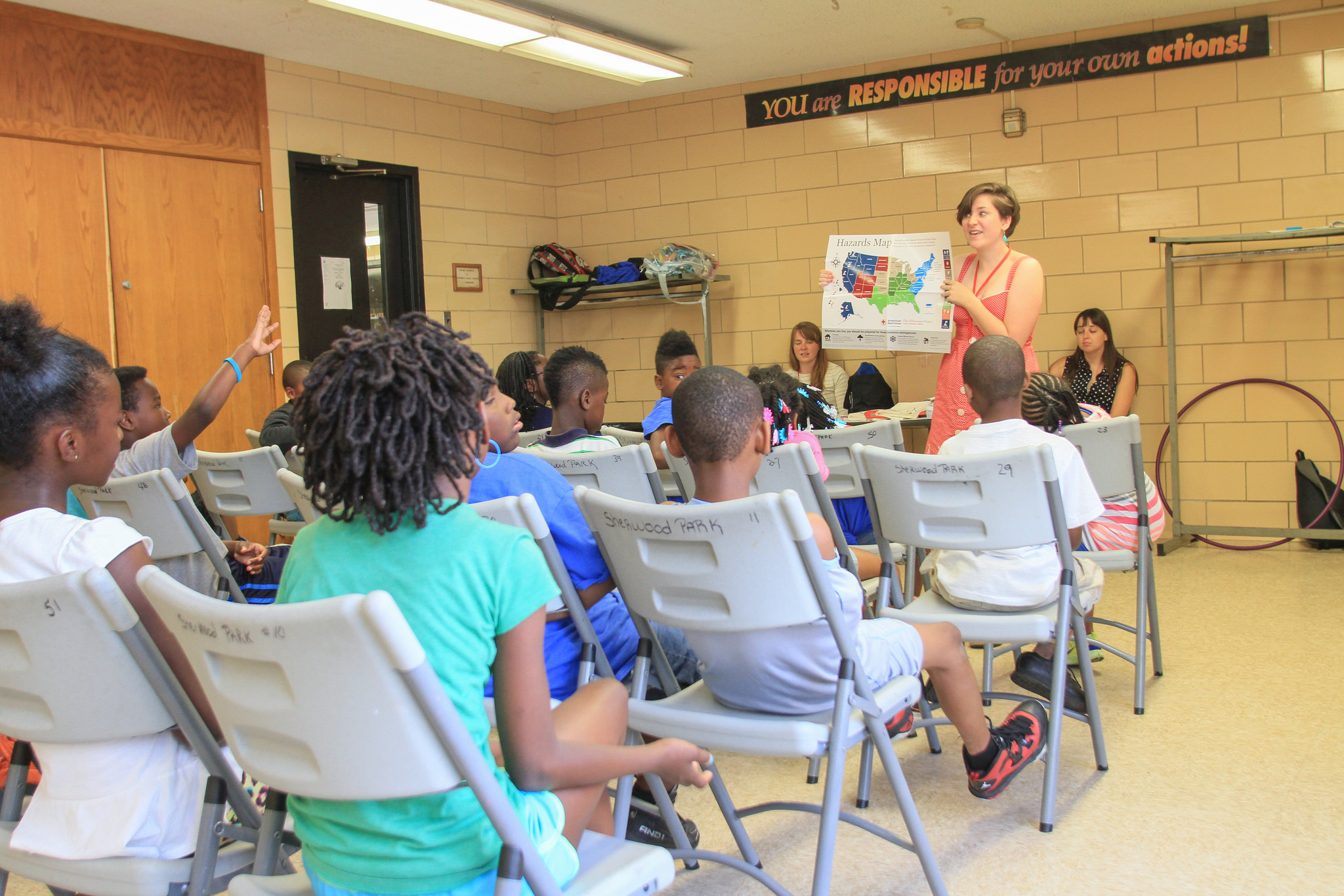 A teacher is standing at the front of a classroom, holding up a hazard map of the United States, while students of diverse backgrounds are seated at desks, listening attentively and raising their hands.