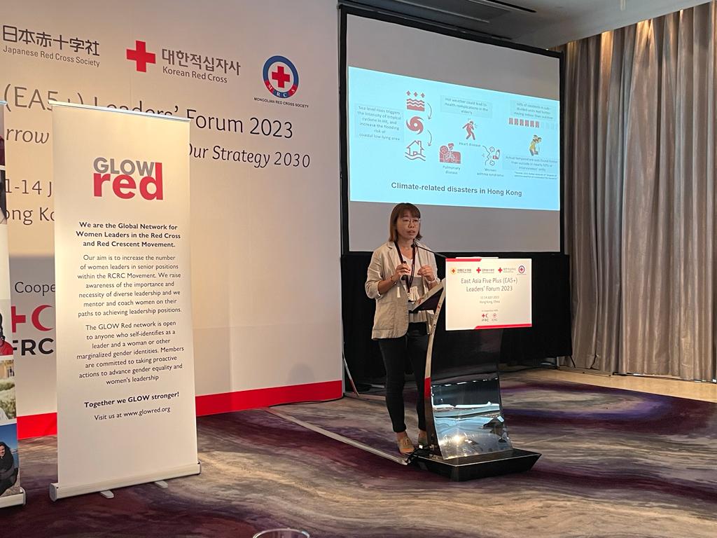 Eva Yeung stands at a lectern in a conference room, delivering a presentation. A slide behind her displays various icons and the text 'Climate-related disasters in Hong Kong'