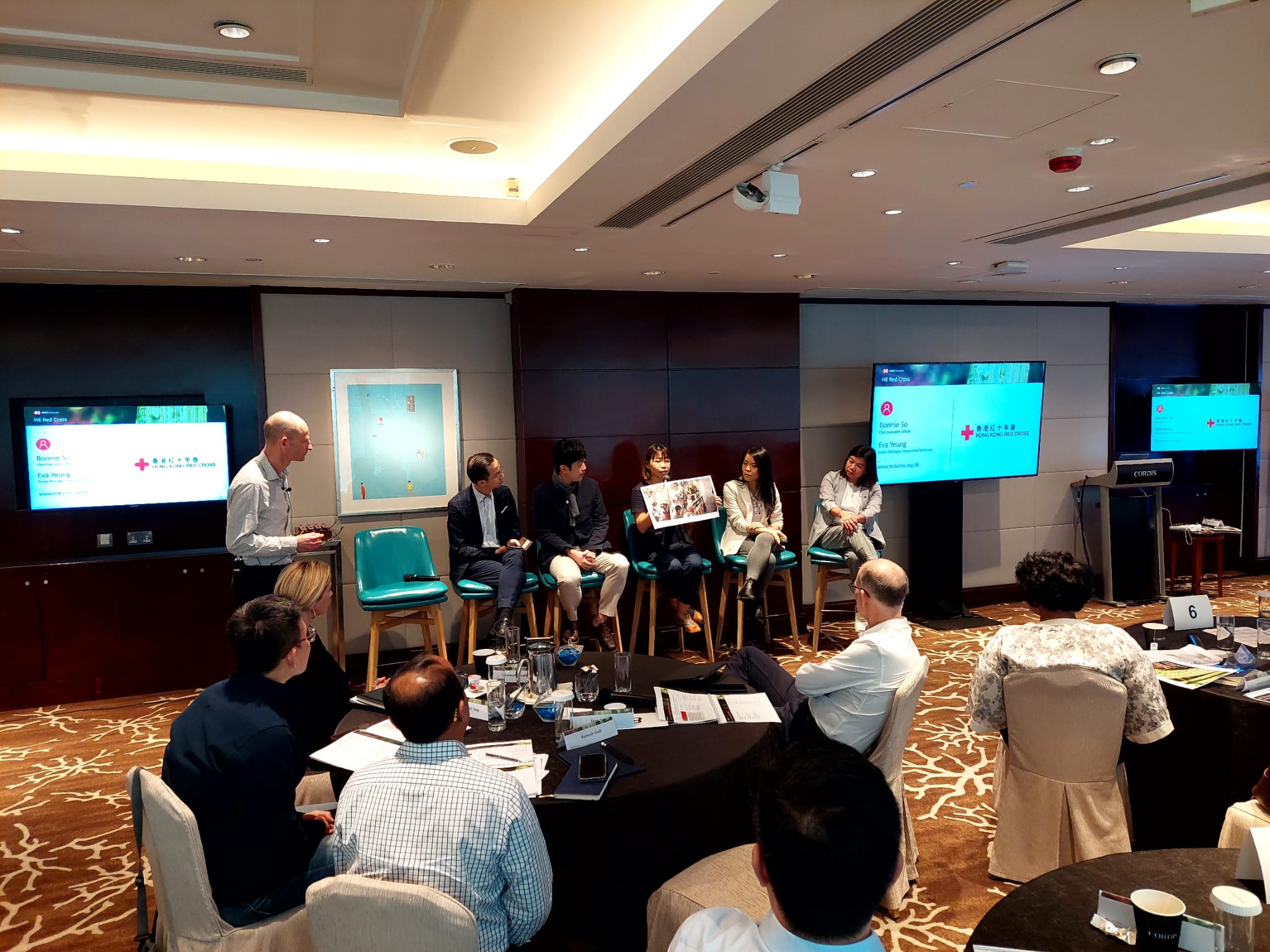 A panel of five individuals sits before an audience in a conference room. Eva Yeung delivers the presentation, holding a banner with photos of sub-divided units in Hong Kong.